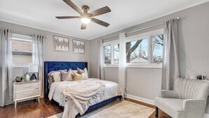 Une chambre à coucher avec un grand lit, un ventilateur de plafond, une table de chevet, une chaise et une fenêtre avec des rideaux.