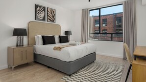 Ein modernes Schlafzimmer mit einem großen Bett, zwei Nachttischen, einem Schreibtisch und Blick auf ein Backsteinhaus durch ein großes Fenster.