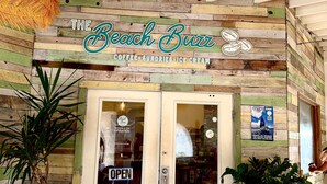 Un café con temática playera, con fachada de madera rústica, un letrero que dice "The Beach Buzz" y una puerta de cristal con un cartel que indica "ABIERTO".