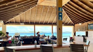 Zona de bar al aire libre con techo de madera y mesas, vistas al mar y un cartel que indica baños de hombres y mujeres.