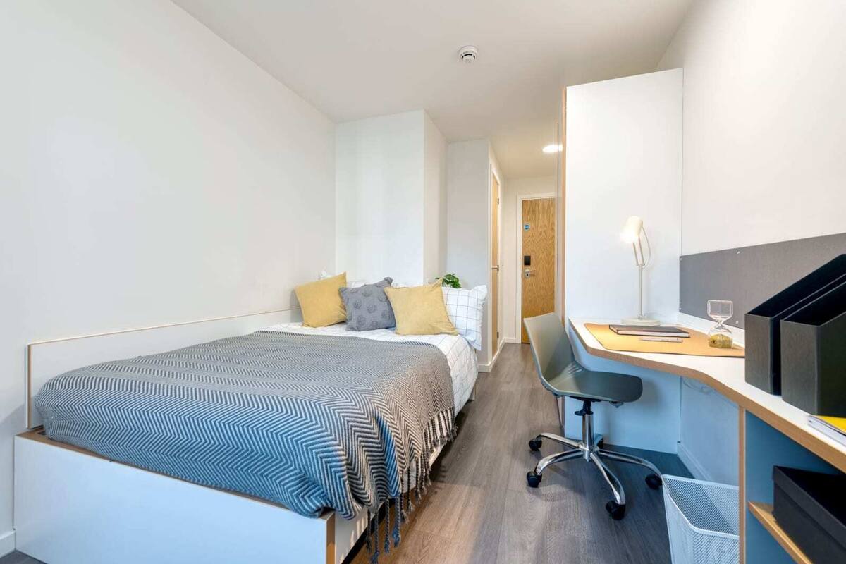 Une chambre à coucher de taille réduite, comprenant un lit, une table de chevet, un bureau, une chaise et une petite poubelle.