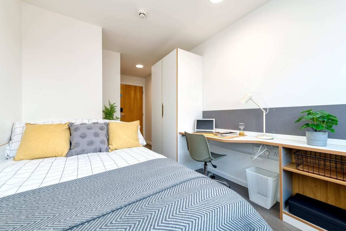 Une chambre moderne avec un lit, un bureau et une chaise. Il y a une plante sur le bureau et une poubelle à côté.