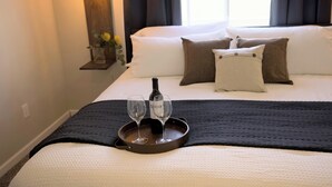 Una cama bien tendida con una bandeja que sostiene una botella y dos vasos, una manta oscura y una mesita de noche con un jarrón de flores.