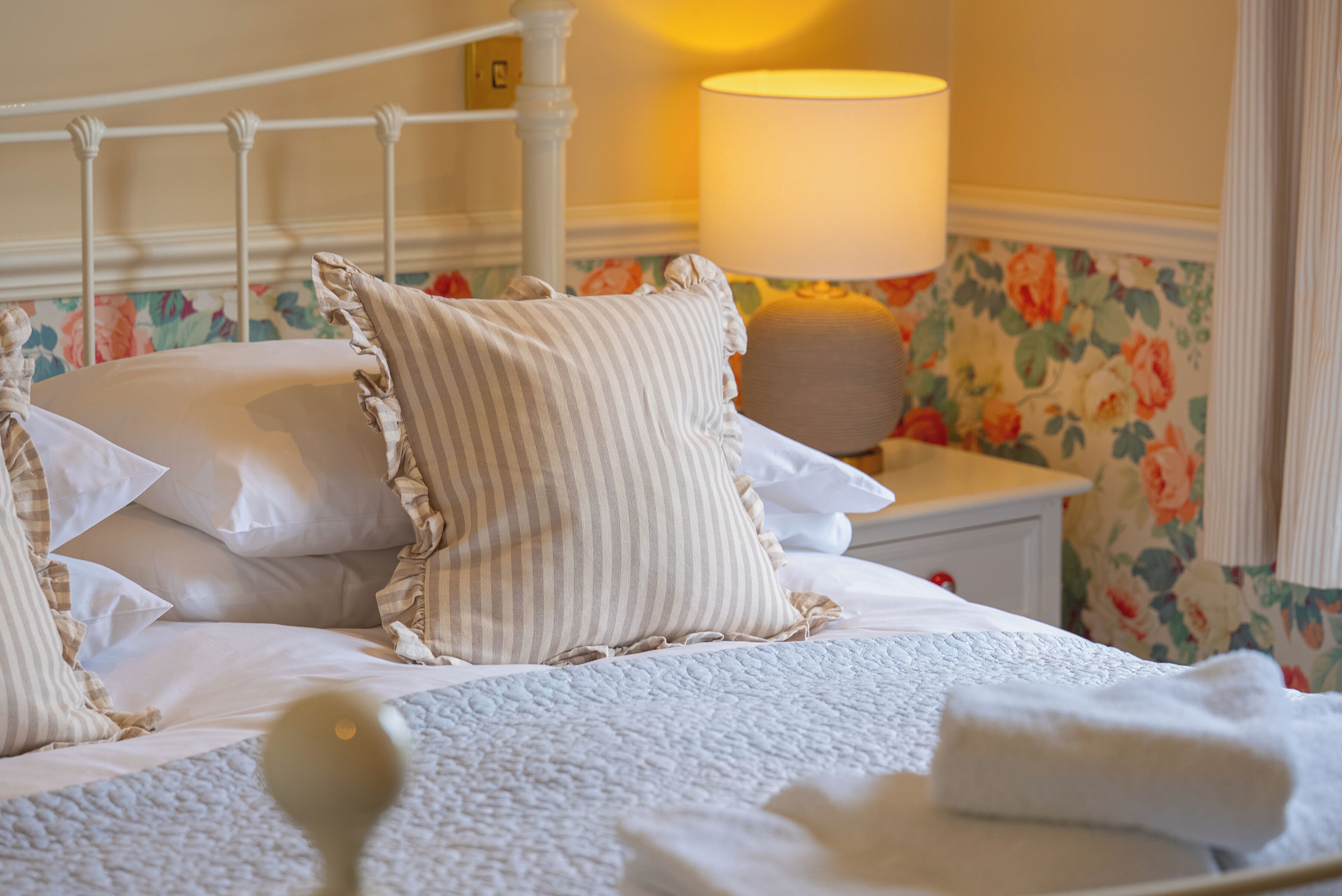 A neatly made bed with pillows and a blanket, a bedside lamp, and a floral wallpaper.