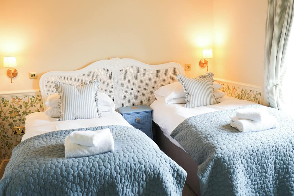 Habitación doble con dos camas, cada una con cubrecama azul acolchado y almohadas blancas. Hay dos mesitas de noche con lámparas, y el papel tapiz de las paredes tiene motivos florales.