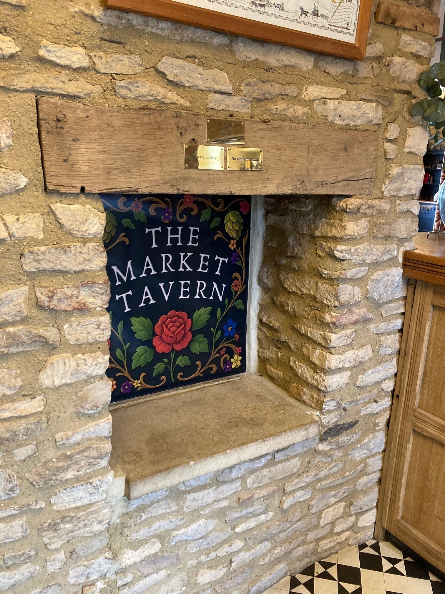 Parede de pedra com uma prateleira de madeira acima de um letreiro que diz 'THE MARKET TAVERN'.