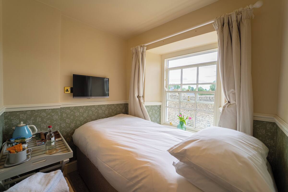 Habitación de hotel con cama, televisor, ventana con cortinas, mesita con tetera y un jarrón con flores.