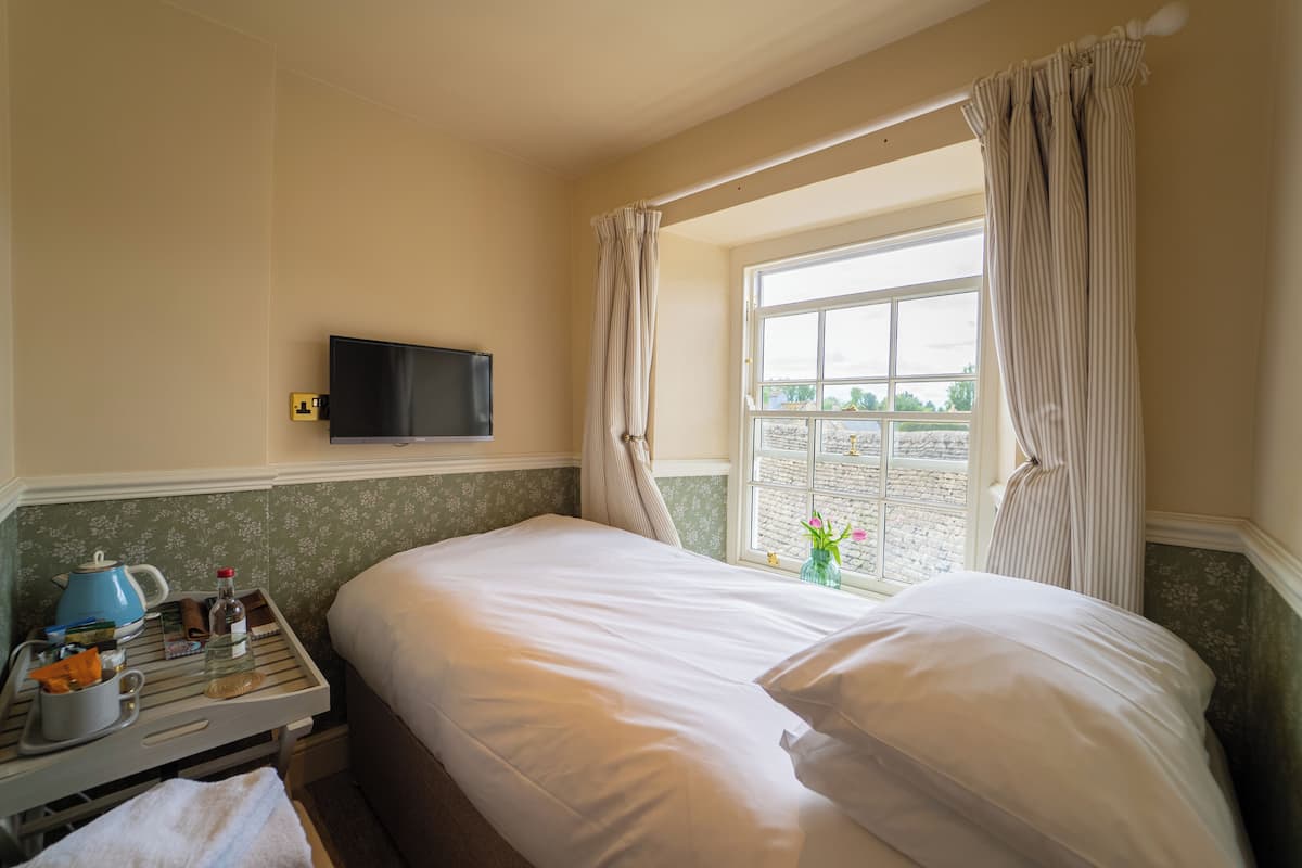 Ett hotellrum med en sÀng, en TV, ett fönster med gardiner, ett litet bord med en vattenkokare och en vas med blommor.