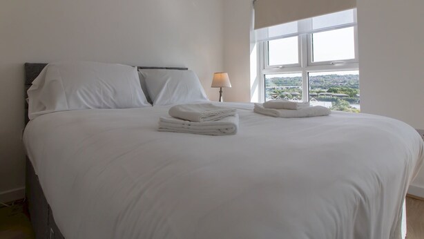 Una cama bien hecha con sábanas y toallas blancas, una lámpara de noche y una ventana con vistas al paisaje.
