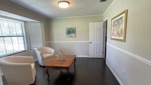 Una habitación con piso de madera, una silla blanca, una mesa de madera con un florero, una puerta y un cuadro en la pared.