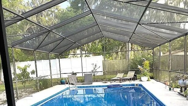 Una piscina rectangular con agua cristalina azul, rodeada por un porche cubierto con sillones de descanso y plantas en macetas.