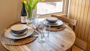 A wooden dining table set with plates, bowls, cutlery, and a wine bottle. There are yellow flowers in a vase, and a window with blinds in the background. - Pass the Keys | Bright and cosy flat in the centre of Much Wenlock (Much Wenlock)