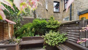 A narrow courtyard with wooden benches, a variety of potted plants, and a brick building in the background. - Stylish Garden Apartment in Vibrant Leyton - Pass the Keys (London)
