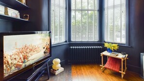 A room with a large flat-screen TV on a stand, a radiator, a wooden table with a vase of flowers, and a bookshelf with various items. - Stylish Garden Apartment in Vibrant Leyton - Pass the Keys (London)