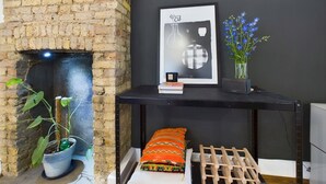 Une pièce avec une cheminée en briques, une console noire surmontée d’un tableau encadré, un vase avec des fleurs bleues et un coussin aux couleurs vives.