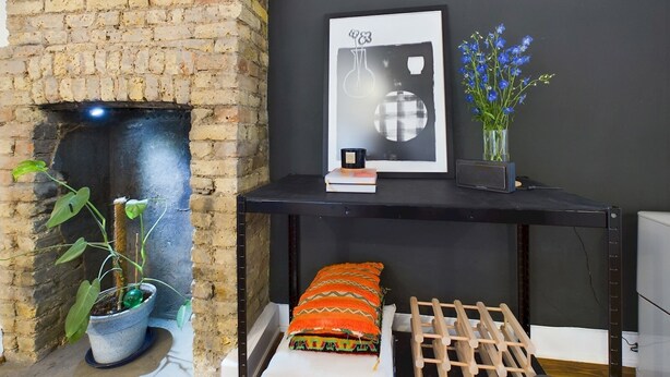 A room with a brick fireplace, a black console table with a framed artwork, a vase with blue flowers, and a colorful cushion. - Stylish Garden Apartment in Vibrant Leyton - Pass the Keys (London)