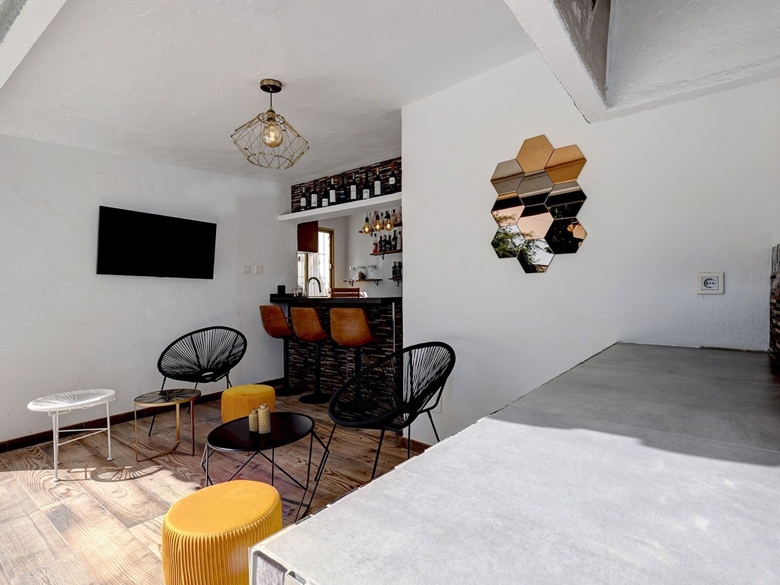 Ein modernes Wohnzimmer mit Barbereich, ausgestattet mit einem Flachbildfernseher, einer einzigartigen sechseckigen Wanddekoration und verschiedenen Sitzgelegenheiten, darunter ein weißer Tisch, gelbe Hocker und schwarze Stühle.