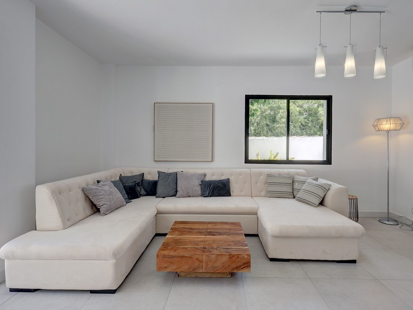 A modern living room with a sectional sofa, a wooden coffee table, pendant lights, a floor lamp, and a window with a view of greenery.