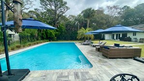 Piscina con sillas de descanso y sombrillas, rodeada de árboles y una casa.
