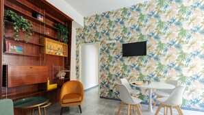 A dining area with a white table and chairs, a wooden bookshelf, and a tropical leaf-patterned wall. - JOIVY Charming 2BR flat near the Duomo (Milan)