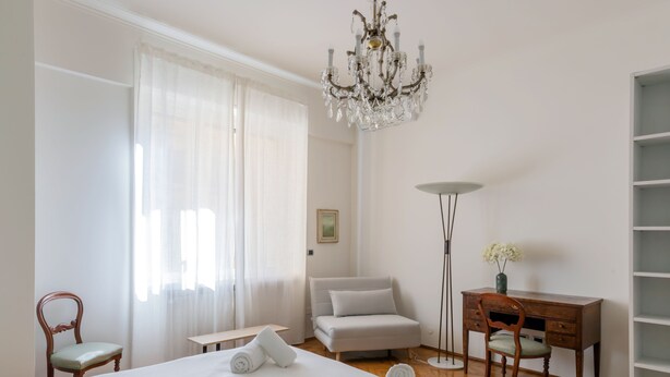 A hotel room with a large bed, a desk, a chair, a floor lamp, and a chandelier. - JOIVY Charming 2BR flat near the Duomo (Milan)