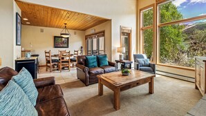 A spacious living room with a wooden ceiling, a large window with a scenic view, a brown leather sofa, a wooden coffee table, and a dining area with a table and chairs. - 4bd Townhome, Mtn Views, Pool & Hot Tub 1079 (Steamboat Springs)