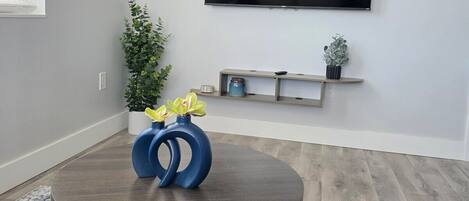 Ett modernt vardagsrum med ett runt bord, en platt-TV som är monterad på väggen, en liten hylla med dekorativa föremål och en krukväxt.