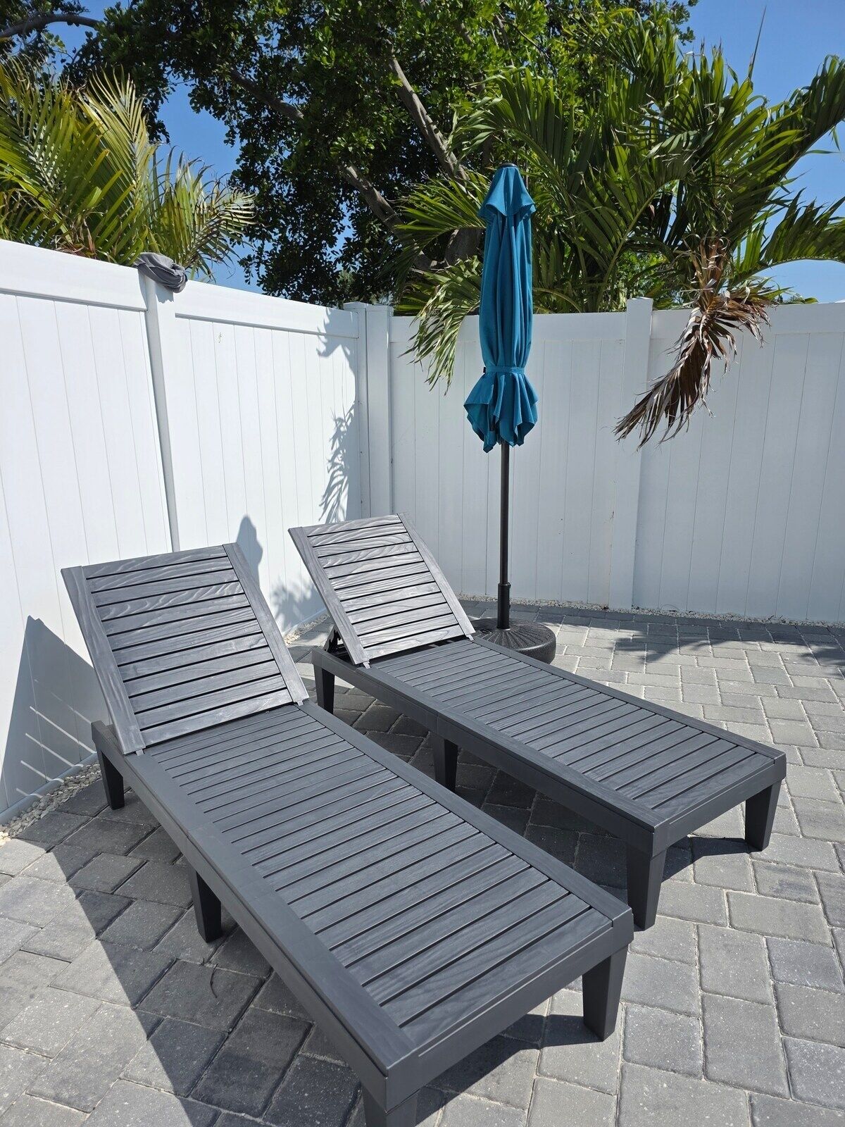 Deux fauteuils de jardin en bois foncé, dotés d’une structure en métal, sont placés sur une terrasse dallée. On aperçoit en arrière-plan une clôture blanche et des palmiers.