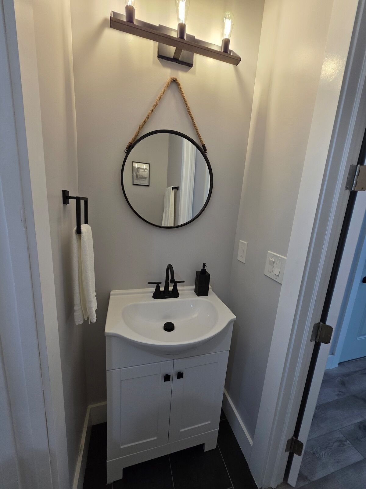 A bathroom with a white sink, black faucet, and a round mirror with a rope handle. There is a towel rack with a white towel and a black soap dispenser on the sink.