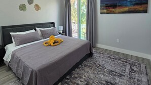 Un dormitorio con cama, ventilador de techo, ventana grande con cortinas, un cuadro en la pared y alfombra estampada.