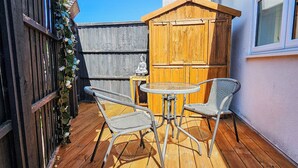 A wooden deck with two wicker chairs and a small table, a wooden fence, and a building with a window.