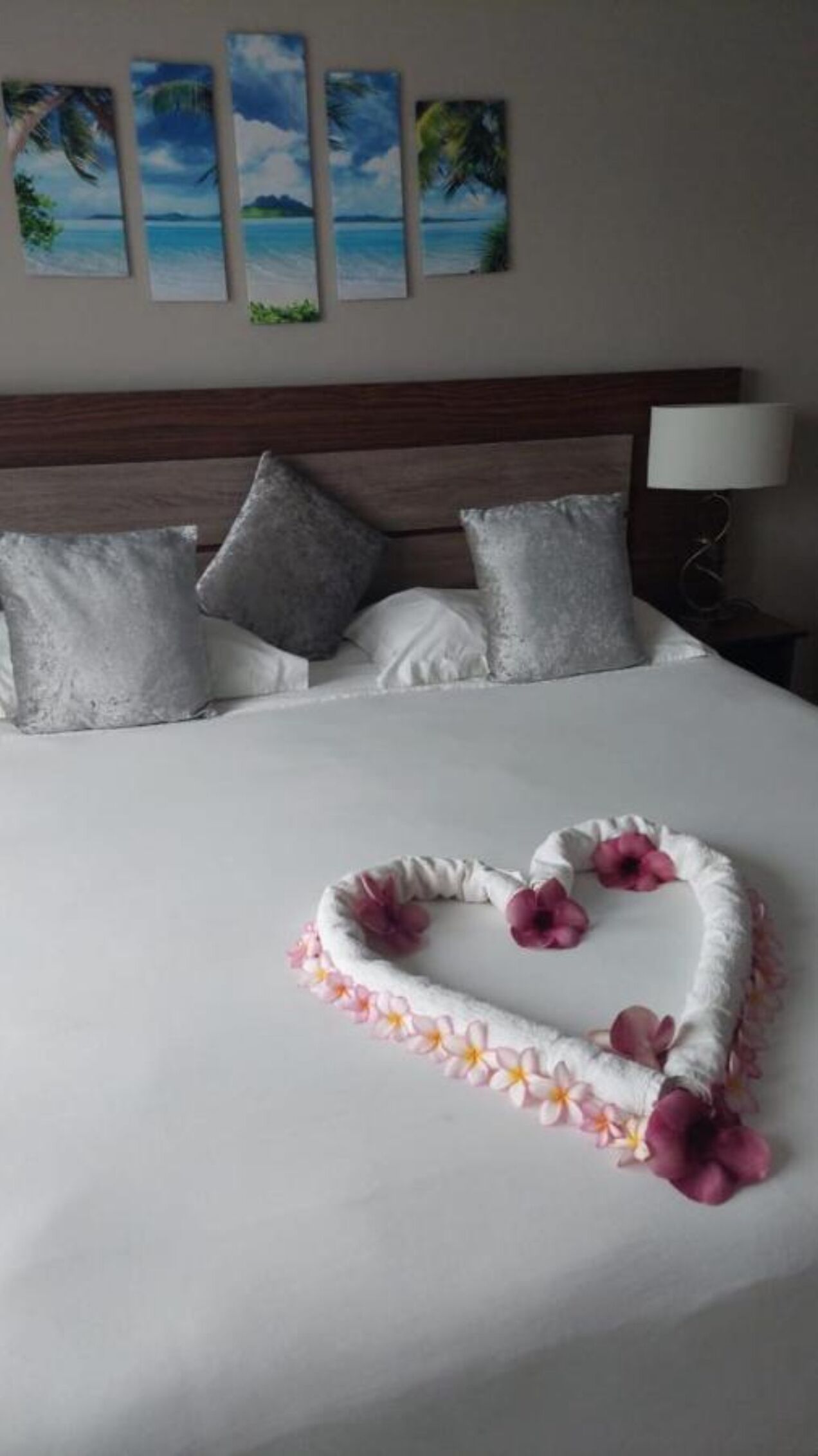 Een net opgemaakt bed met een bloemstuk in hartvorm in het midden, een houten hoofdbord en een muur met vier ingelijste schilderijen van tropische stranden.