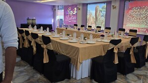 Ein Konferenzraum mit langem Tisch, gedeckt für eine Besprechung, mit schwarzen Stühlen, die mit beige farbenen Bändern geschmückt sind, und einem Banner mit der Aufschrift 'Ladies Kitty Party' an der Wand.