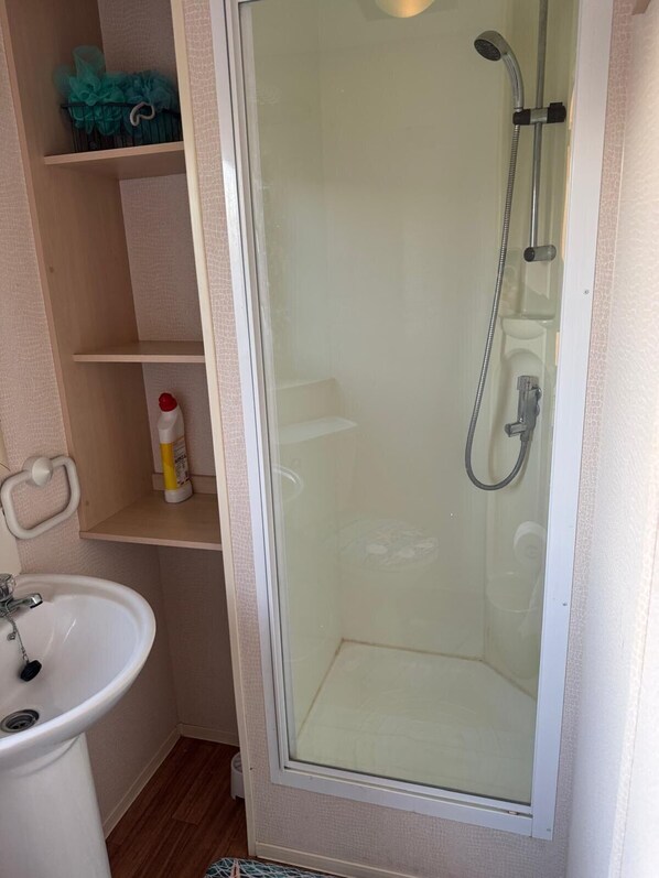 Un baño con ducha de mampara de vidrio, un lavabo y estantes con artículos de aseo.