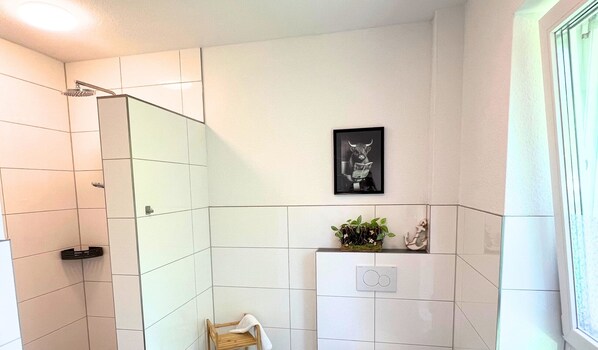 A bathroom with white tiles, a shower, a sink with a mirror, and a framed picture on the wall. - Apartment "Neptun" Papenburg 2SZ EBK Modern bathroom Large roof terrace New building WLAN (Papenburg)
