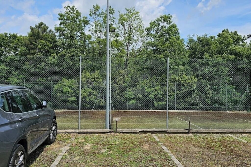 A fenced sports court with a parked car and trees in the background.