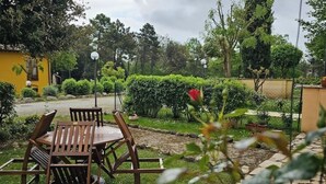 An outdoor seating area with a wooden table and chairs, surrounded by greenery and a yellow building in the background.