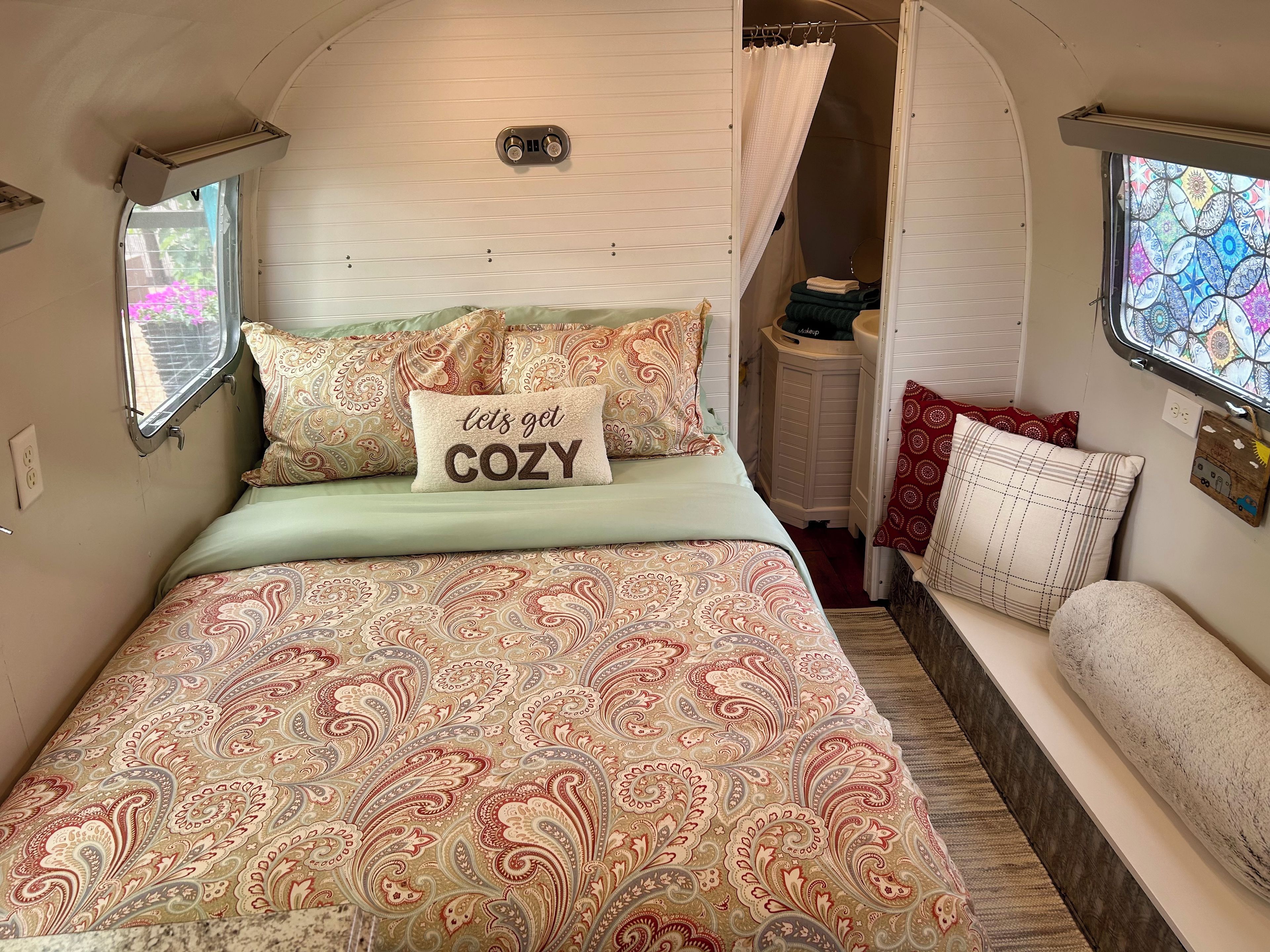 A cozy interior with a bed featuring a 'let's get COZY' pillow, patterned bedding, and a small window with a view.