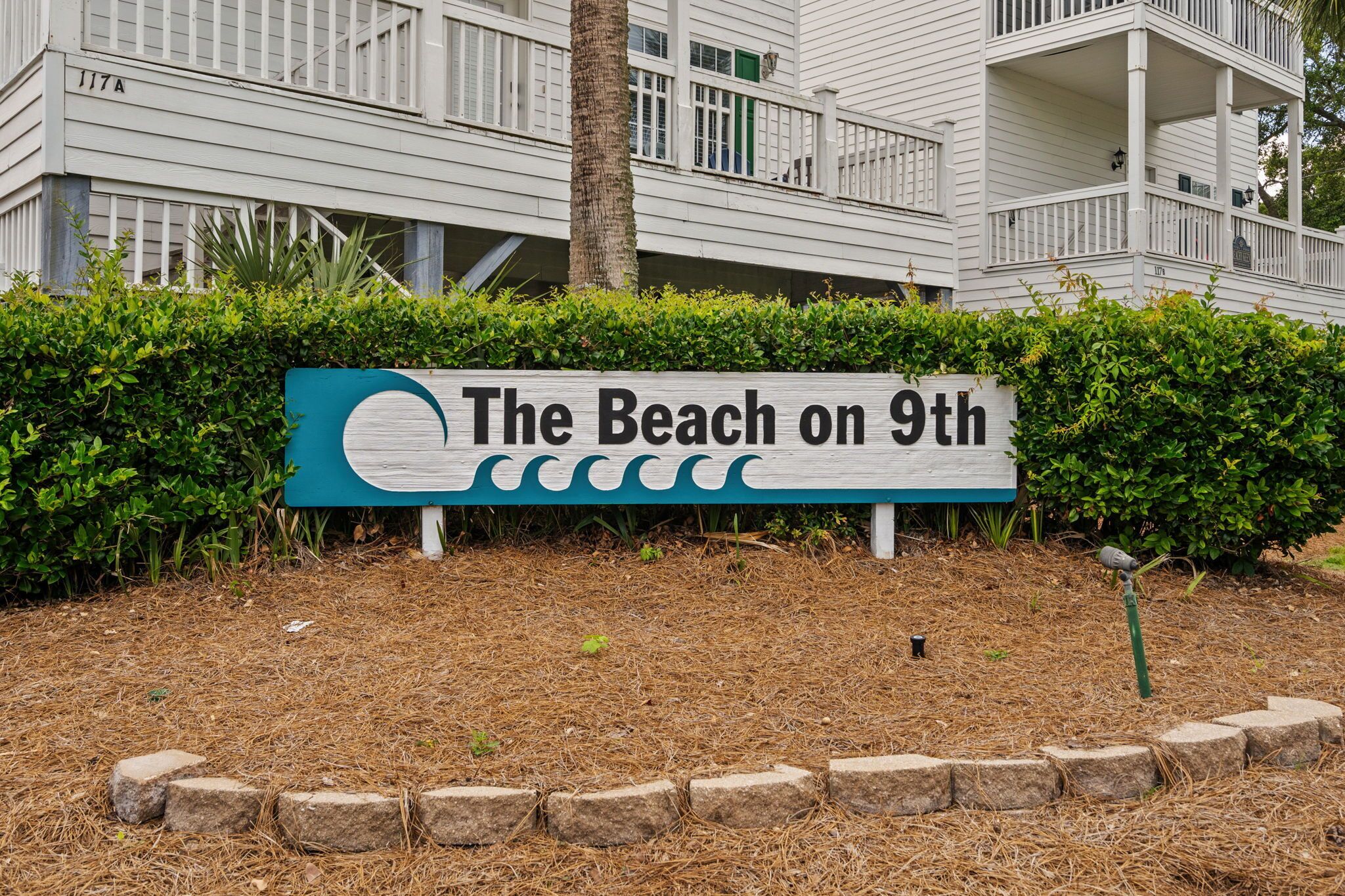 Letrero que dice 'The Beach on 9th', con un diseño de ola, frente a un edificio blanco con balcones y un seto.