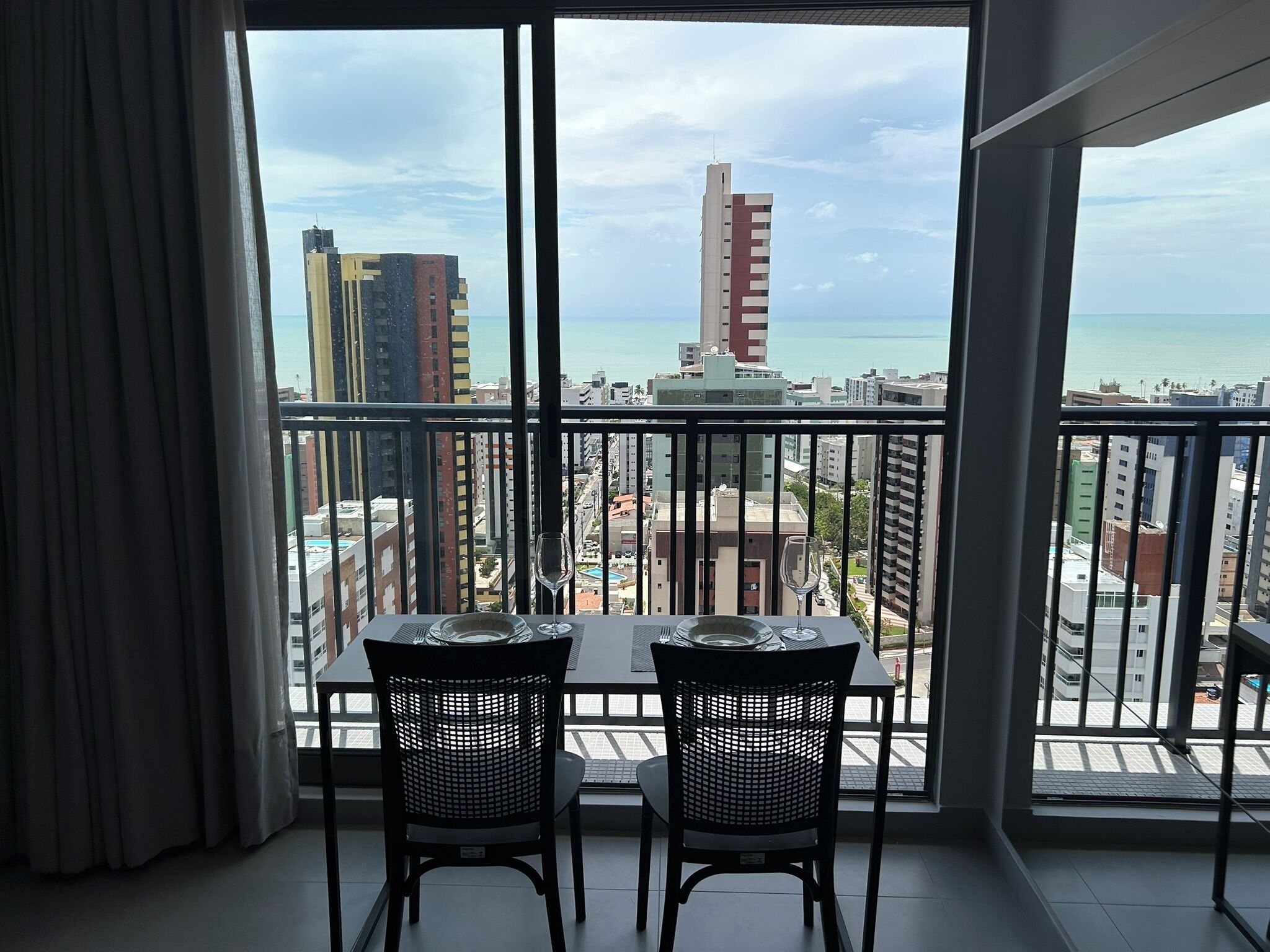 A balcony with a table and chairs, overlooking a cityscape and the sea.