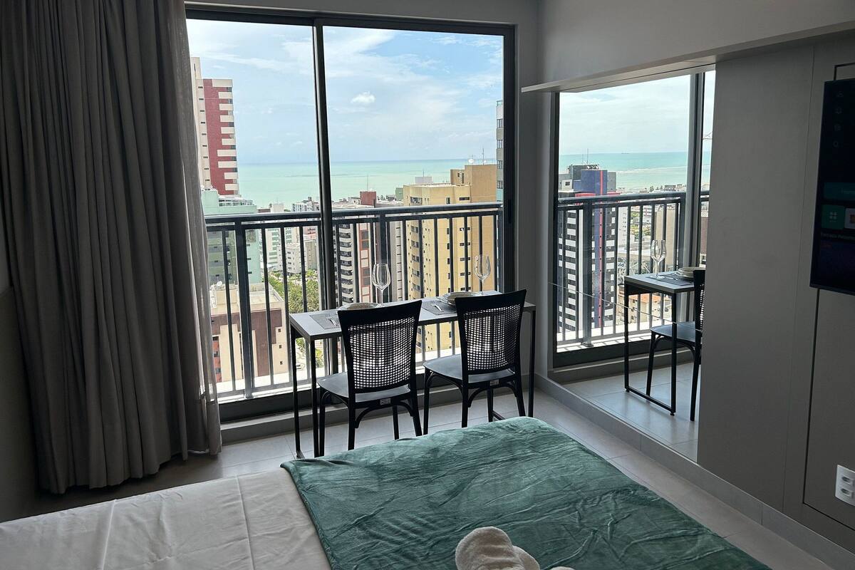A hotel room with a balcony, a bed, a green throw, and a view of the city and sea.