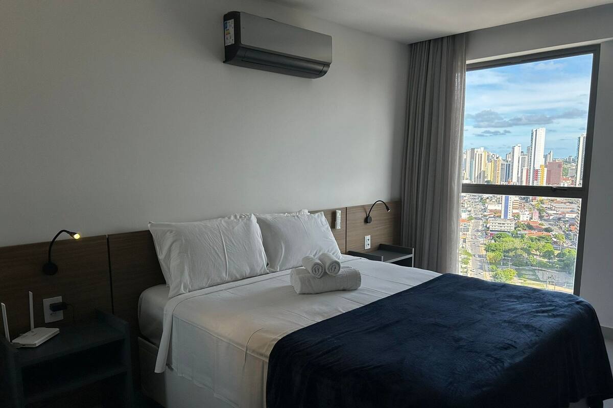 A hotel room with a bed, a nightstand, a lamp, an air conditioner, and a window with a city view.