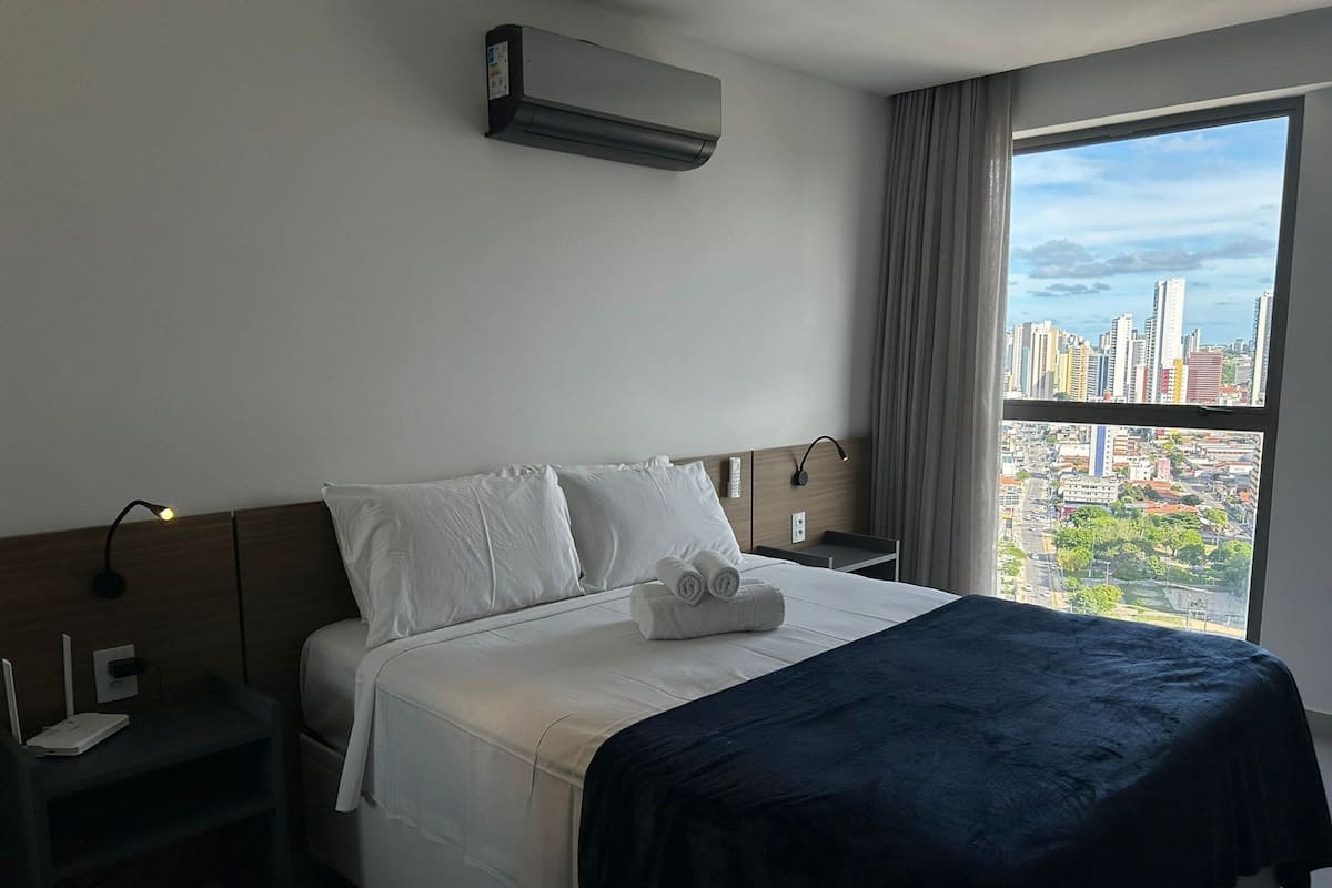 Hotellihuone, jossa on sänky, yöpöytä, lamppu, ilmastointilaite ja ikkuna, josta on näkymä kaupunkiin.