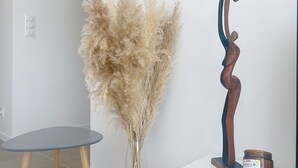 A room with a round table, a vase with pampas grass, a wooden sculpture, and a candle on a white pedestal.