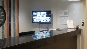A reception desk with a flat-screen TV displaying multiple security camera feeds, a clock, and a welcome sign. - Home Place Suites (Bartlesville)