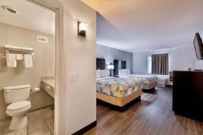 A hotel room with a toilet, two beds with patterned bedspreads, a bedside table, a lamp, and a large mirror.