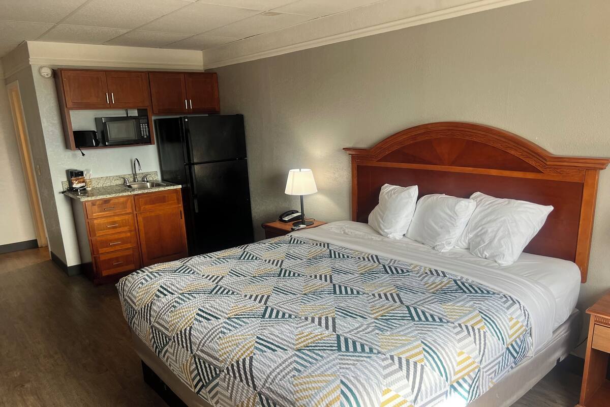 A hotel room with a bed, a nightstand, a lamp, a wooden headboard, a kitchenette with a refrigerator, microwave, and cabinets.