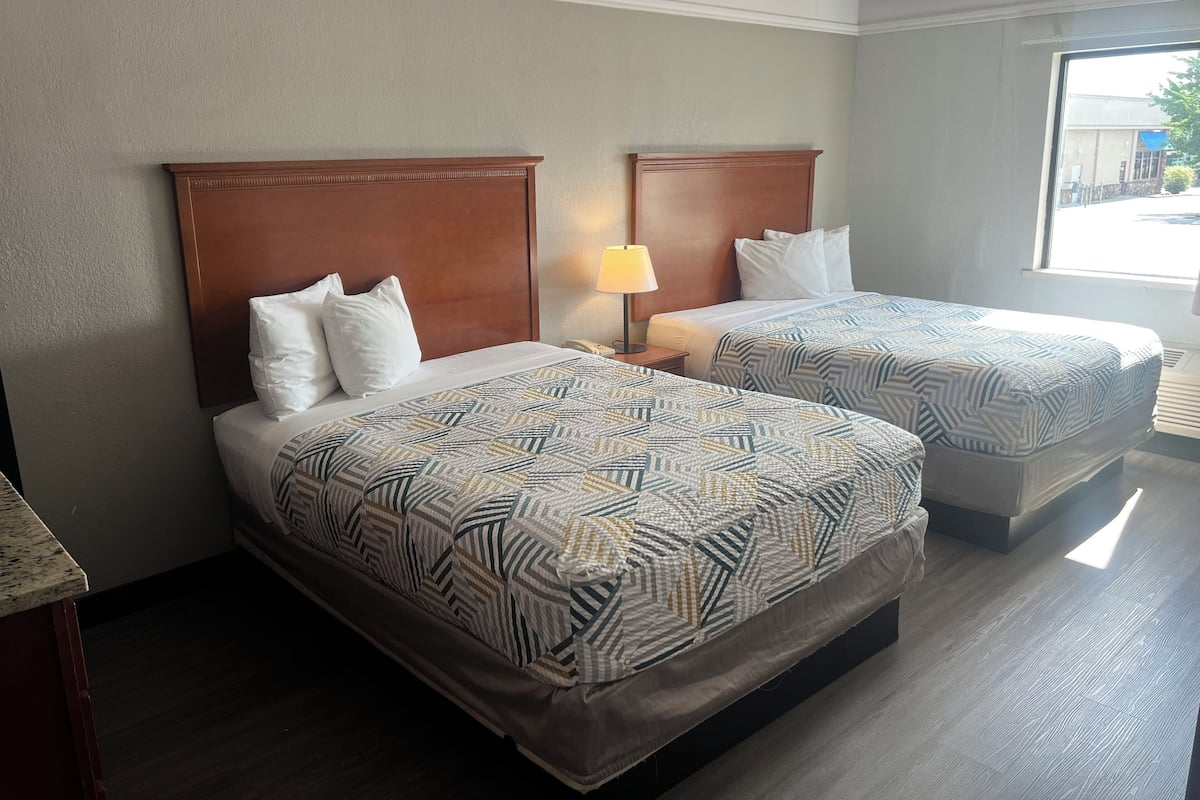 A hotel room with two beds, each with a patterned bedspread and white pillows. There is a nightstand with a lamp between the beds.