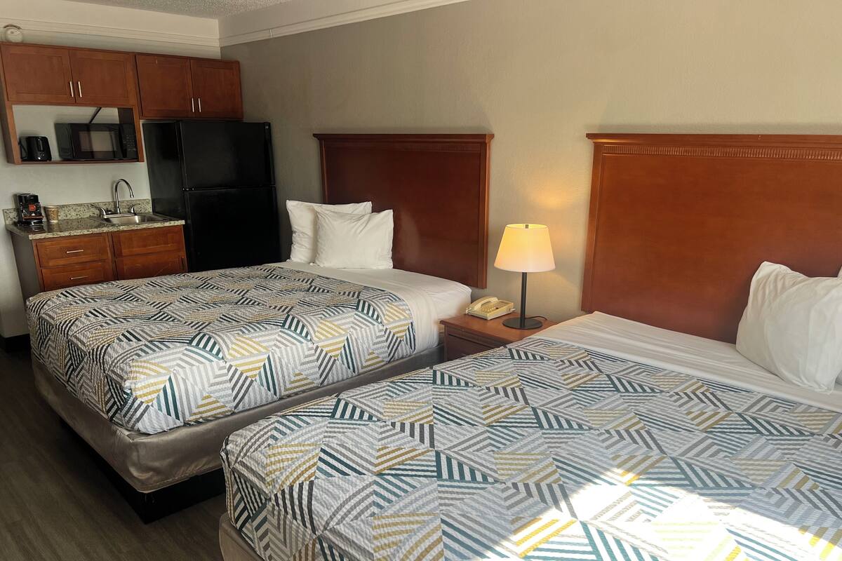 A hotel room with two beds, a kitchenette, and a bedside lamp.