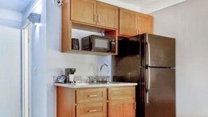 A compact kitchen with wooden cabinets, a stainless steel refrigerator, and a microwave on the countertop. - Home Place Suites (Bartlesville)